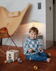 A toddler in a blue checkered sweater and jeans sits barefoot on a tiled floor, playing with wooden toys and the mushie Silicone Pacifier Clip | Hera. A modern butterfly chair and small lamp add to the cozy, minimalist design.