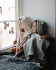 Two kids in mushie Bear Poncho Towels smile by a window, wearing cozy organic cotton.