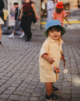 A toddler in yellow holds toys and a mushie Linen Pacifier Clip 2-Pack on a cobblestone street.