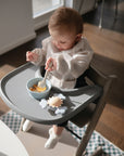 A baby in a high chair uses the mushie Dinnerware Cutlery Set—non-toxic, dishwasher-safe plastic—with a light blue bowl, sunlight streaming in and a crab-shaped toy on the tray.