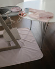 A wooden high chair with a pink mat sits by a white table holding a pink bib, divided plate, pink pacifier on a chain, and a mushie Dinnerware Cup in blue. The dark wood floor enhances the cozy atmosphere.