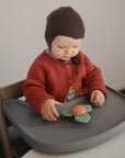 A baby in a high chair plays with the mushie Suction Spinner Toy, a green and orange flower-shaped sensory toy made of food-grade silicone for safe play.