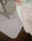 A Mushie Splat Mat in pale pink is under a wooden chair beside a white table with a pink cloth and pastel baby toy, on a dark wood floor.