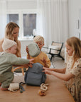 A woman and three children pack a light blue backpack at a softly lit, neutral-toned table. The baby reaches for the bag as the others help, holding mushie brand Lunchboxes made of non-toxic plastic.