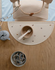 Overhead view of a mushie baby feeding set on a wooden table: rainbow-print silicone placemat, beige bowl and spoon, grey bear pacifier clip, and a small bowl filled with mushie Chain Link Rings for fine motor skills.