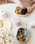 A child’s hand reaches for grapes in a divided bowl of fruit on a white table, surrounded by mushie Round Dinnerware Plates, baby cups, a plate with fruit slices, a bowl of nuts and kiwi, and a vase of daisies.