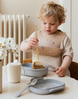 A young child with curly blond hair enjoys cake from a mushie Lunchbox at the table, wearing a beige bib with small figures. On the table are a white sippy cup and flowers.