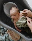 A baby is wrapped in a mushie Organic Cotton Muslin Cloths 3-Pack while lying in a gray baby bathtub. An adult gently brushes the baby's arm, with towels visible nearby.