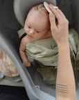 An adult gently bathes a baby wrapped in a green towel, with mushie Organic Cotton Muslin Cloths 3-Pack nearby for nursery needs. The baby lies in a small tub, looking up as the adult’s tattooed forearm is visible.