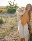 A smiling woman carries a happy child wrapped in a mushie Organic Cotton Muslin Swaddle Blanket.