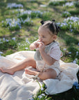 A baby sits on a mushie Organic Cotton Muslin Swaddle Blanket outdoors, surrounded by small blue flowers and green grass, wearing a light outfit and sucking their fingers while sunlight filters through the scene.