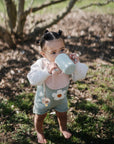 A young child with small buns stands barefoot on grass, wearing a daisy overall and sipping from a mug outdoors near a tree—enjoying the moment among baby essentials like the FRIGG Moon Natural Rubber Pacifier 2-Pack by FRIGG.