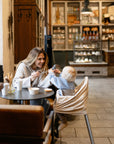 A woman smiles at a baby in a high chair, playfully holding her glasses as they interact with mushie Chain Link Rings—colorful toys that help develop fine motor skills. Shelves with food and drinks create a cozy café backdrop.