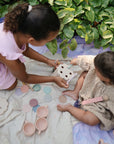Two children play on a mushie Organic Cotton Muslin Swaddle Blanket near green foliage.