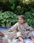A smiling toddler in a beige dress enjoys a picnic outdoors, surrounded by green plants and her favorite FRIGG Daisy Night Natural Rubber Pacifier 2-Pack.