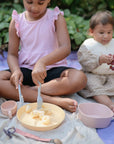 Two young children enjoy a picnic on a blanket outdoors, using the mushie Dinnerware Cutlery Set. One slices bananas while the other holds grapes, surrounded by cups, a teapot, and bowls with greenery in the background.