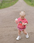 Toddler with mushie Linen Pacifier Clip 2-Pack in pink outfit holding a snack on dirt road.