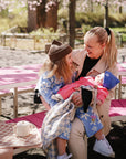 A woman holds two kids with mushie Linen Pacifier Clip 2-Pack at a pink picnic table.