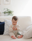 A baby in a cream knit outfit sits on a white couch with pillows and a cozy blanket, playing with a beaded teether. In the background is the FRIGG Butterfly Anatomical Silicone Pacifier 2-Pack (6-18 Months) by FRIGG.