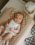 Smiling baby with mushie Linen Pacifier Clip 2-Pack on a checkered changing pad, diapers nearby.