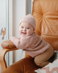 A smiling baby in a pink knit hat and sweater enjoys a mushie Suction Spinner Toy on a tan leather chair. A colorful cushion and window with flower decals appear in the background as the baby looks at the camera.