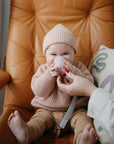 A smiling baby in a pink knit outfit sits on a tan leather chair, holding a pink cup with help from an adult. Beside the baby is a patterned pillow and the mushie Linen Pacifier Clip 2-Pack.