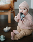 A baby in a knit hat and sweater sits on a wooden floor, chewing on a mushie Bath Boat. Two more mushie Bath Boats are nearby, with a chair in the background.