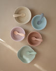 Five mushie Silicone Toddler Starter Spoons, each paired with a pastel bowl in cream, blue, pink, peach, and mint green, rest on a beige surface. Soft lighting enhances their gentle colors—ideal for encouraging toddlers to self-feed.