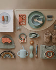 A flat lay of baby items in soft pastels, including a bib, mushie Silicone Toddler Starter Spoons for self-feeding, teethers, a cup, crocheted booties and beanie, a sweater, rainbow stacker, toy car, and boxed silicone toothbrushes.