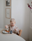 A young child eats a meal with rice and sauce at a round table, using a spoon. Their food is served on a mushie Silicone Placemat in a light, minimalist room with framed art and red leaves on the windowsill.