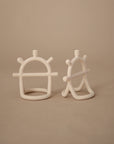Two abstract, white No-Drop Sensory Teethers by mushie, with curved frames and short protruding rods, stand on a neutral beige surface against a plain beige background.