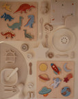 A flat lay of children’s toys features dinosaur and space-themed wooden puzzles, a silicone bib, utensils, a white star, a rainbow, the Mushie Classic Silicone Suction Plate for toddlers, a soft space cloth, and textured non-toxic plates on beige.