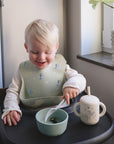 A smiling blonde toddler sits in a high chair, wearing a mushie Bluey x Mushie Silicone Baby Bib, eating from a bowl with a spoon. Next to the bowl is a straw cup. Sunlight shines in from a window with a potted plant nearby.