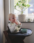 A young blond child sits in a high chair by a window, wearing a bib and holding the mushie Bluey x Mushie Snack Cup. A green bowl and spoon are on the tray with other baby feeding essentials, and a potted plant is on the windowsill behind.