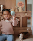 A young child sits in a wooden playroom using a mushie Linen Pacifier Clip 2-Pack, with a pacifier attached. They're holding a round toy, while a toy monkey and wooden bowls rest on the bench beside them.