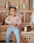 A young child with pigtails, in a pink sweater and light blue pants, smiles while sitting on a wooden play kitchen surrounded by wooden toys, utensils, and the mushie Sealife Mold Free Bath Play Set.