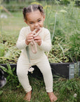 A young child in a cream outfit sits barefoot on a garden bed edge, smiling and chewing on a FRIGG Daisy Natural Rubber Pacifier. Green plants grow behind them, and part of a black dog is visible in the foreground.