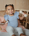A young child with pigtails sits on a wicker couch, smiling and holding a pale rattle, a toy, and a mushie Linen Pacifier Clip 2-Pack with silicone clasp. The child wears a light blue top and white leggings with a checkered cushion behind.