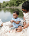 A woman and baby play with mushie Nesting Stars Toy on a blanket by a lake.