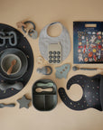 A flat lay of mushie baby items in soft blue, gray, and beige includes bibs, Silicone Toddler Starter Spoons for self-feeding, bowls, a teether, a divided lunchbox, and an outer space-themed sticker sheet on a beige backdrop.