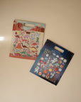 Two packs of mushie sticker sets on a light beige surface: one with dinosaur puffy stickers against a desert background, and the other is a reusable space-themed sticker set featuring astronauts, planets, and rockets on a starry backdrop.