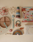 A flat lay of baby items in soft pastels, featuring a divided silicone plate, silicone utensils, stacking toys, teether, pacifier, rainbow toy, muslin cloth, soft textured toy, and the mushie Reusable Sticker Set.