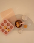 A flat lay shows Mushie’s BPA-free Baby Food Freezer Tray in pink with round food portions, alongside a gray placemat, brown bowl with pink spoon, and a white baby food storage container featuring a shaker top.