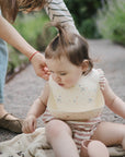 Woman adjusts bib on baby outdoors, using mushie Bluey Gift Set on a blanket.
