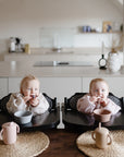 Two babies sit side by side in high chairs at a kitchen table, each holding a teething toy. In front of them are mushie Silicone Toddler Starter Spoons, bowls, and sippy cups for independent feeding, with a modern kitchen in the background.