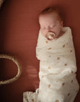Newborn wrapped in mushie Organic Cotton Muslin Swaddle Blanket, lying in a basket bassinet.