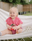 Smiling toddler wears mushie Linen Pacifier Clip 2-Pack on a white hammock outdoors.