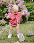 A toddler plays with a Mushie Bath Rinse Cup among lilac and purple flowers.