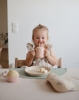 A young blonde child sits at a table, smiling and drinking from a pink straw cup. In front of her are food and a cupcake served on mushie Round Dinnerware Plates, which are non-toxic, dishwasher safe, and perfect for a bright minimal setting.