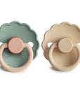 The FRIGG Daisy Natural Rubber Pacifier 2-Pack includes two daisy-shaped pacifiers—one sage green with a pink handle, one cream with a beige handle—both featuring the FRIGG logo and soothing natural rubber nipples.